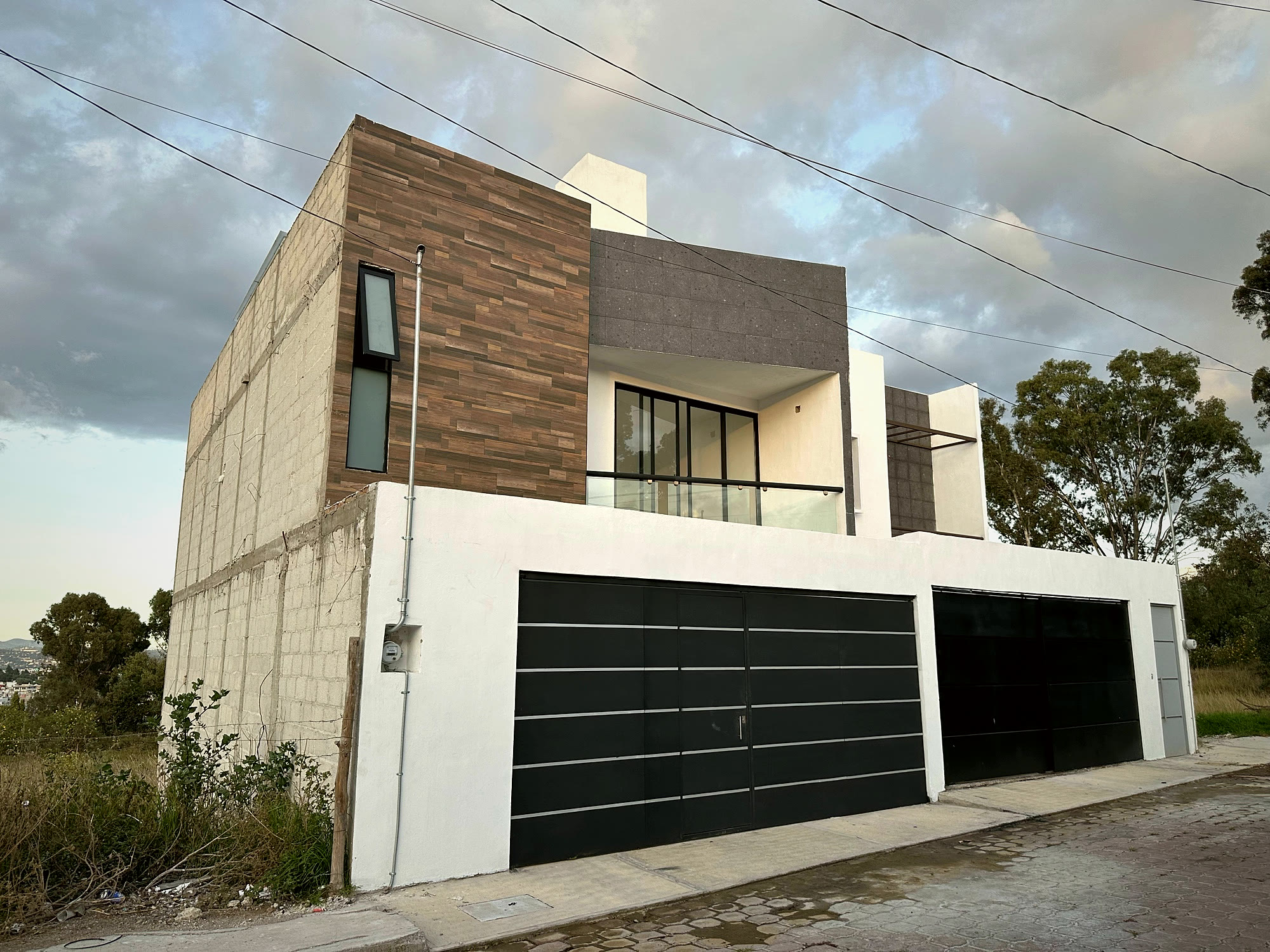 Fachada de las dos casas al atardecer, con balcón de vidrio y caja de madera en la esquina.